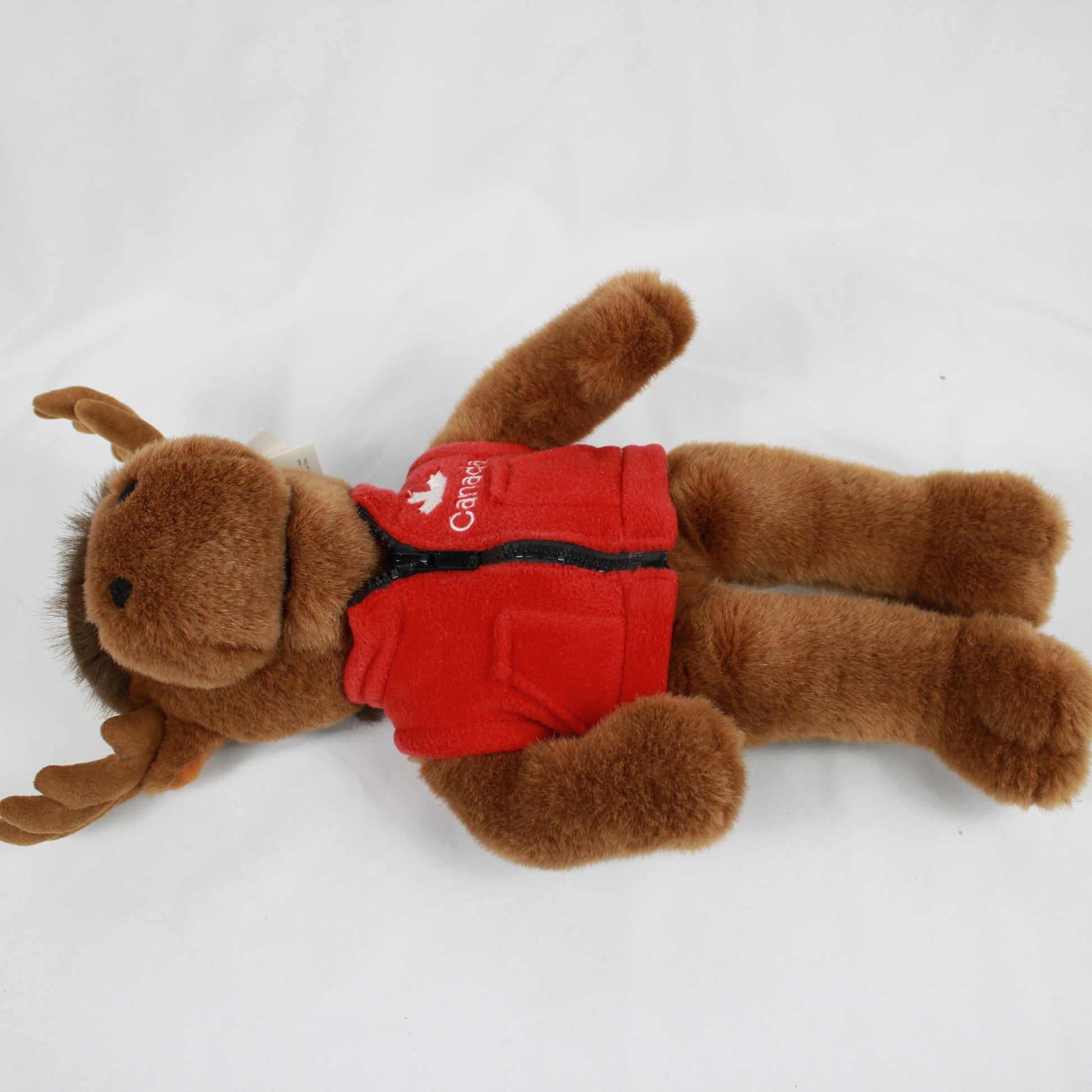 Plush moose toy wearing a red vest and hat with a Canadian flag, on a white background.