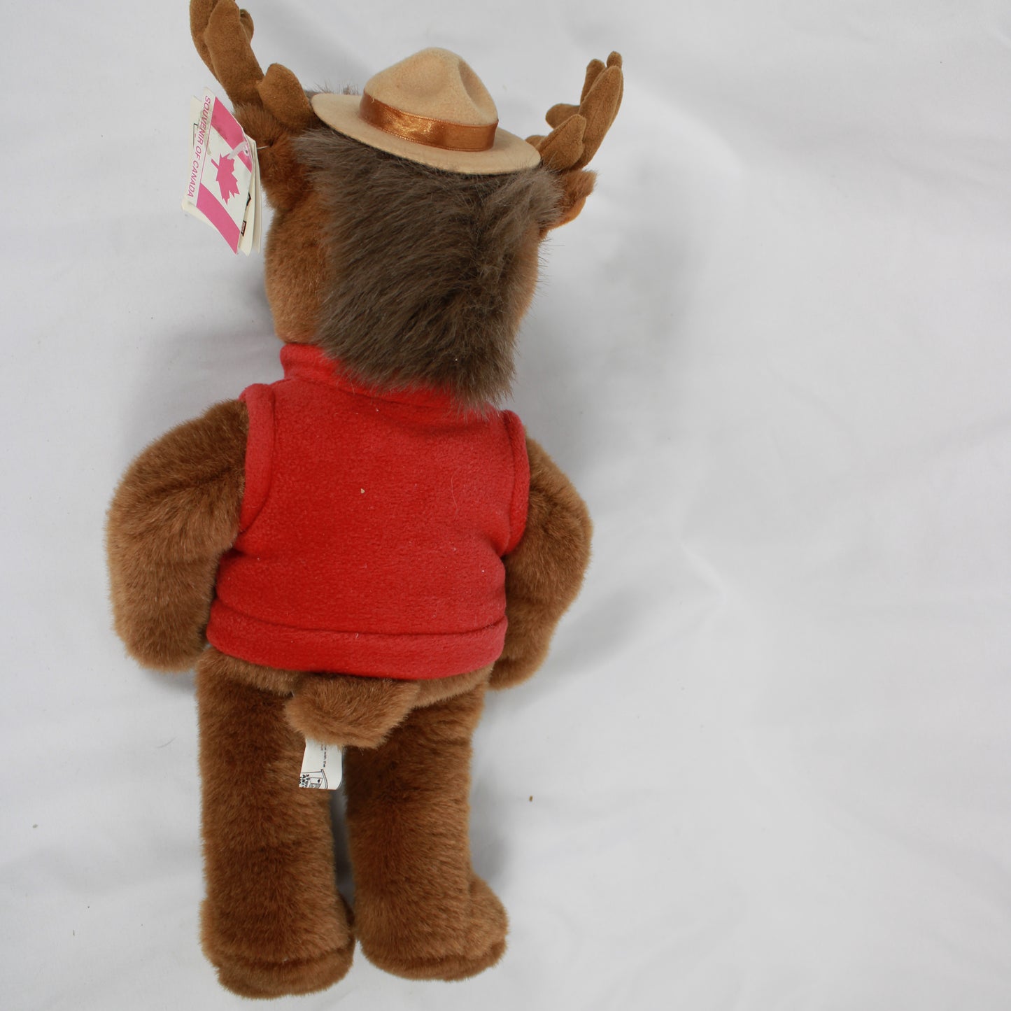 Plush moose toy wearing a red vest and hat with a Canadian flag, on a white background.