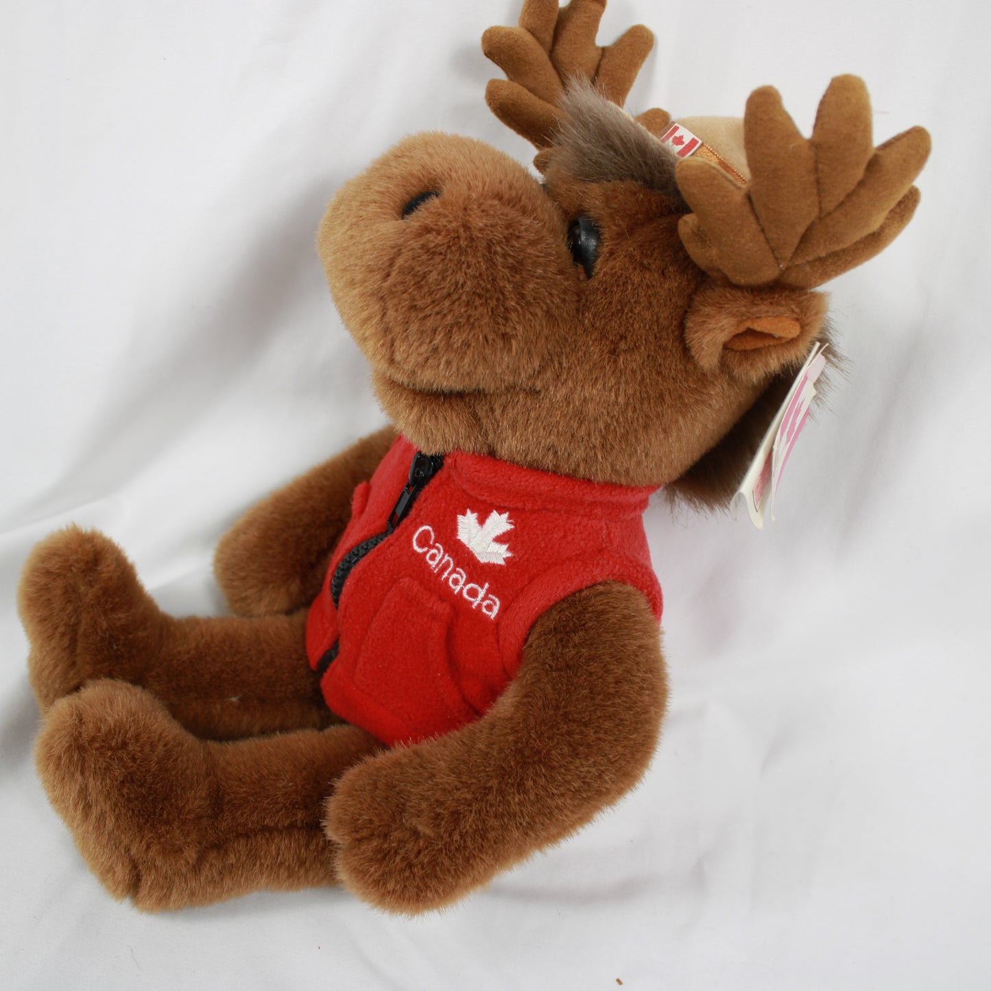 Plush moose toy wearing a red vest and hat with a Canadian flag, on a white background.