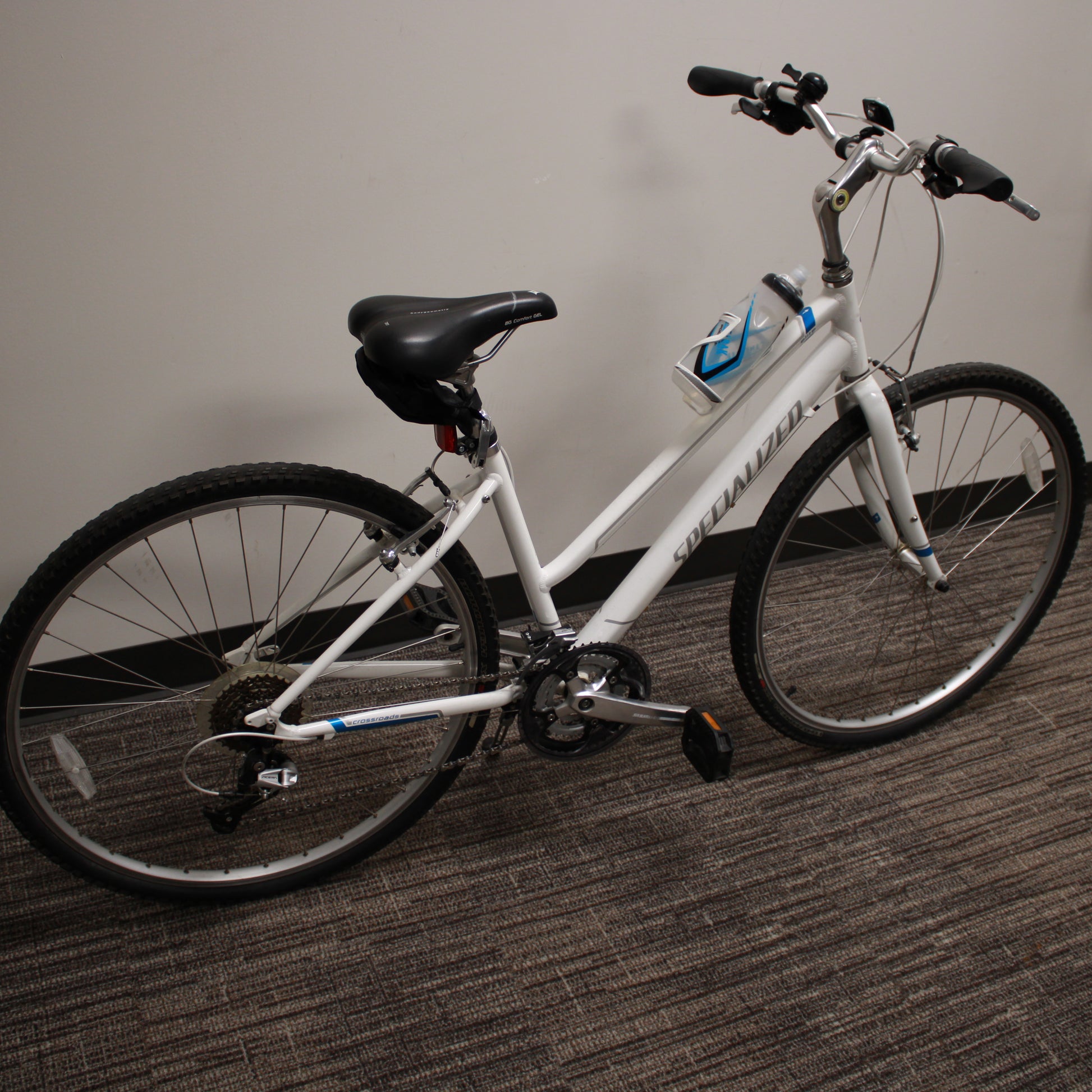 White bicycle with black seat and tires on a carpeted floor against a plain wall.