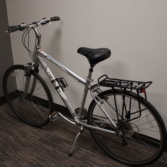 Silver bicycle with a black seat and basket against a plain wall.